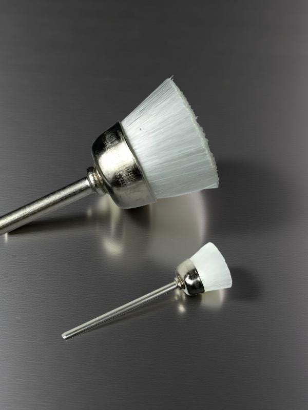 Two small soft nylon brushes with white bristles on metallic bases on a reflective gray surface. These Brush E-File Bit, designed to replace traditional nail brushes for a faster, more efficient cleaning process. 